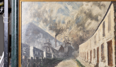 Colliery Row by Norman Cornish in a black frame with a golden edge. The paining portrays a row of terraces houses on one side, with minework buildings opposite, with smoke coming out of a large industrial chimney and the smoke filling the sky.