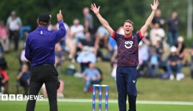 A Lancashire player in short-sleeved blue top with red facings and blue trousers appeals fgor a wciket while the umpire in bright blue shirt and black trousers wit h a black cap raises his right index finger to signify a dismissal. The stumps are blue with sponsor logo. A blurred shot of spectators sitting on a grass verge can be seen in the back ground.
