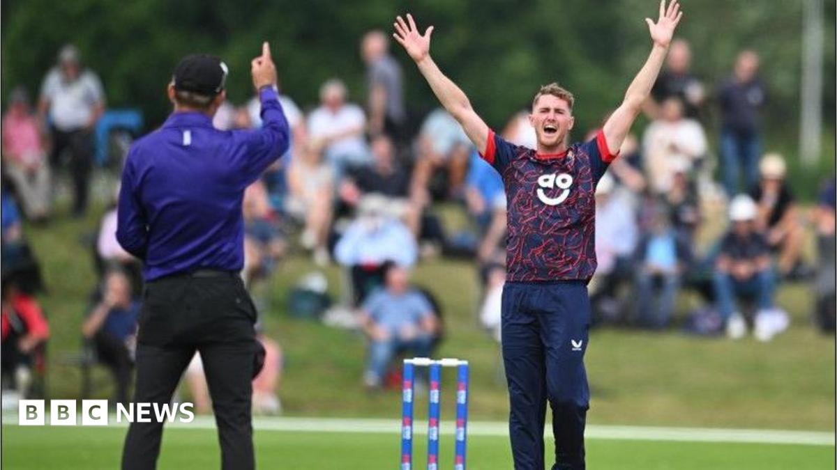 A Lancashire player in short-sleeved blue top with red facings and blue trousers appeals fgor a wciket while the umpire in bright blue shirt and black trousers wit h a black cap raises his right index finger to signify a dismissal. The stumps are blue with sponsor logo. A blurred shot of spectators sitting on a grass verge can be seen in the back ground.