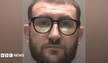 A police mugshot of a man in his early 30s, who has short brown hair and a brown beard. He is wearing black framed glasses and is looking into the camera with a bemused expression.