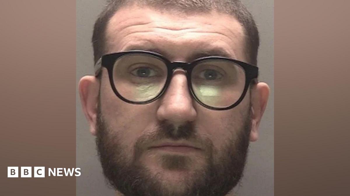 A police mugshot of a man in his early 30s, who has short brown hair and a brown beard. He is wearing black framed glasses and is looking into the camera with a bemused expression.