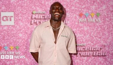 A man is pulling a big smile with his mouth open and is wearing a low-buttoned light pink shirt with some dainty white necklaces. Behind him is a pink wall which has the Garnier logo on it.