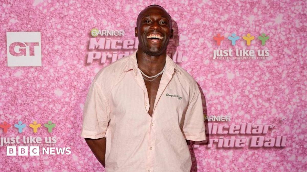 A man is pulling a big smile with his mouth open and is wearing a low-buttoned light pink shirt with some dainty white necklaces. Behind him is a pink wall which has the Garnier logo on it.