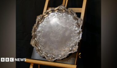 A large circular silver salver is rested on an upright wooden frame. It has a decorative finish.