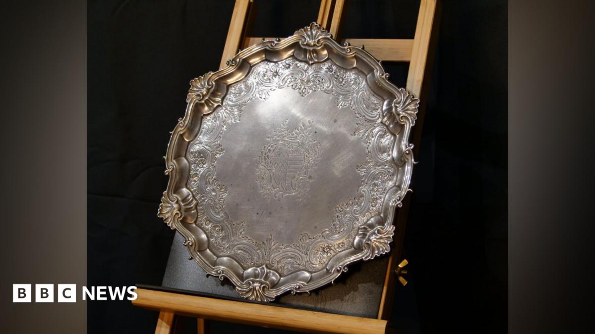 A large circular silver salver is rested on an upright wooden frame. It has a decorative finish.