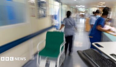 A stock image of NHS workers in a hospital corridor. They are wearing blue scrubs. One is pulling a wheelchair while two are looking at paperwork on a reception desk.