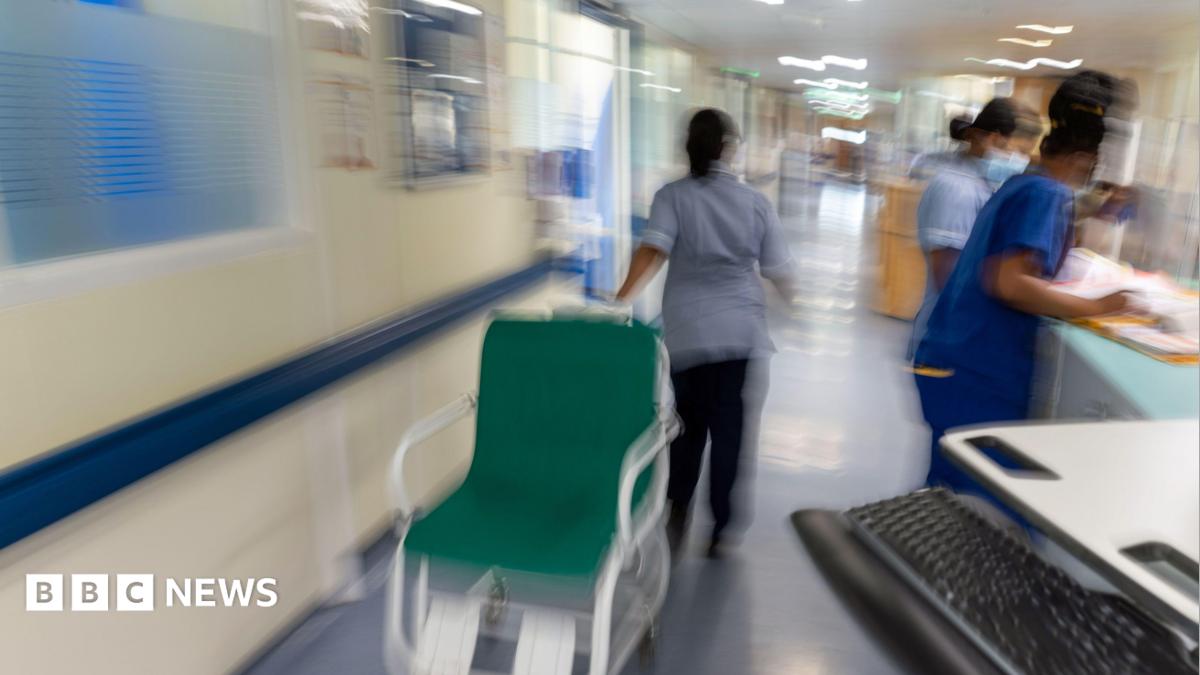A stock image of NHS workers in a hospital corridor. They are wearing blue scrubs. One is pulling a wheelchair while two are looking at paperwork on a reception desk.