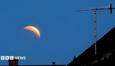 The image shows the crest of a moon against an early morning sky. The roof of a house and a chimney can be seen in the foreground.