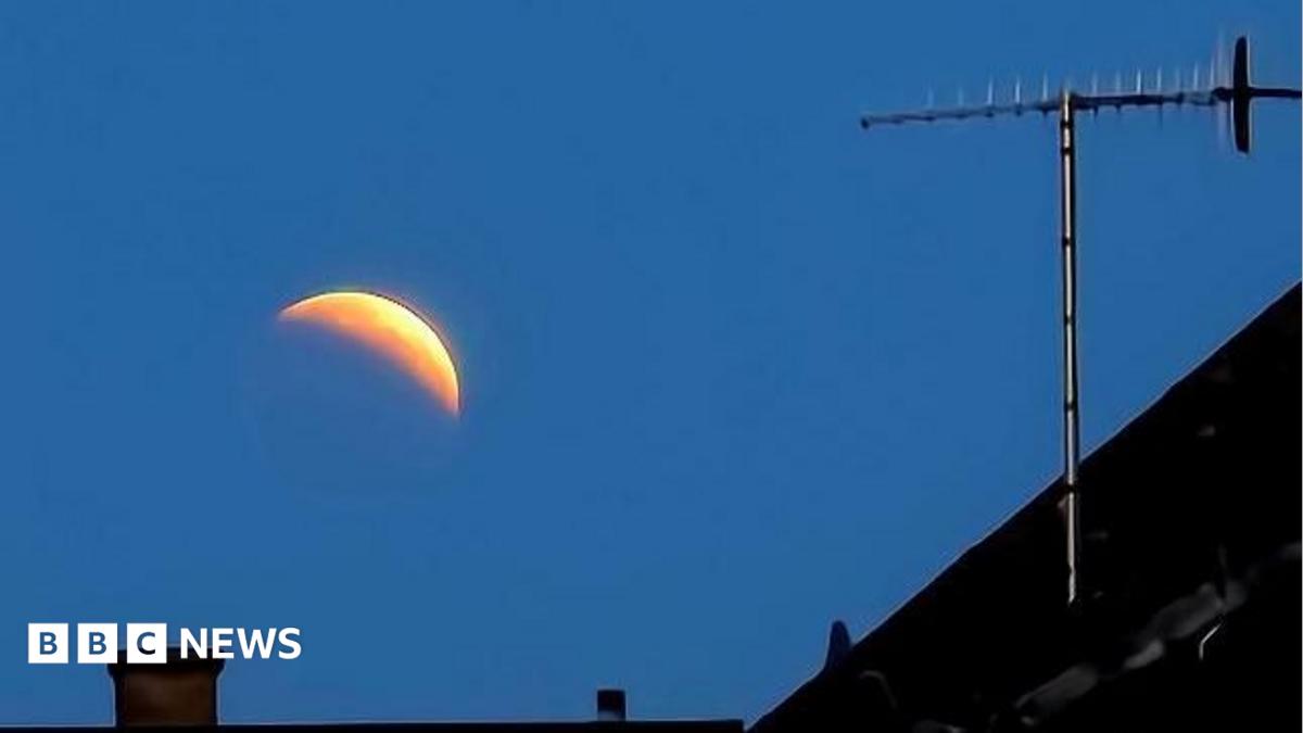 The image shows the crest of a moon against an early morning sky. The roof of a house and a chimney can be seen in the foreground.
