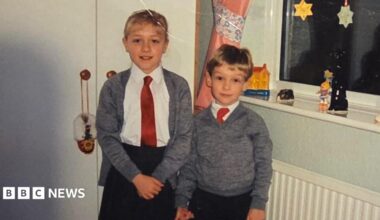 The childhood photo of Claire and Peter which was sent to Dalibor Sentic in the care package he recieved in 1992. Claire and Peter are photographed in school uniforms, stood in what appears to be a bedroom. Claire is taller than Peter and stood to the left of the image. She is wearing a black skirt, white blouse, red tie, and grey cardigan. She has blonde hair with a fringe. Her hair is tied up. Peter is shorter than Claire and is photographed to the right of her. He is wearing black trousers, a white shirt and red tie, with a grey jumper over the top.