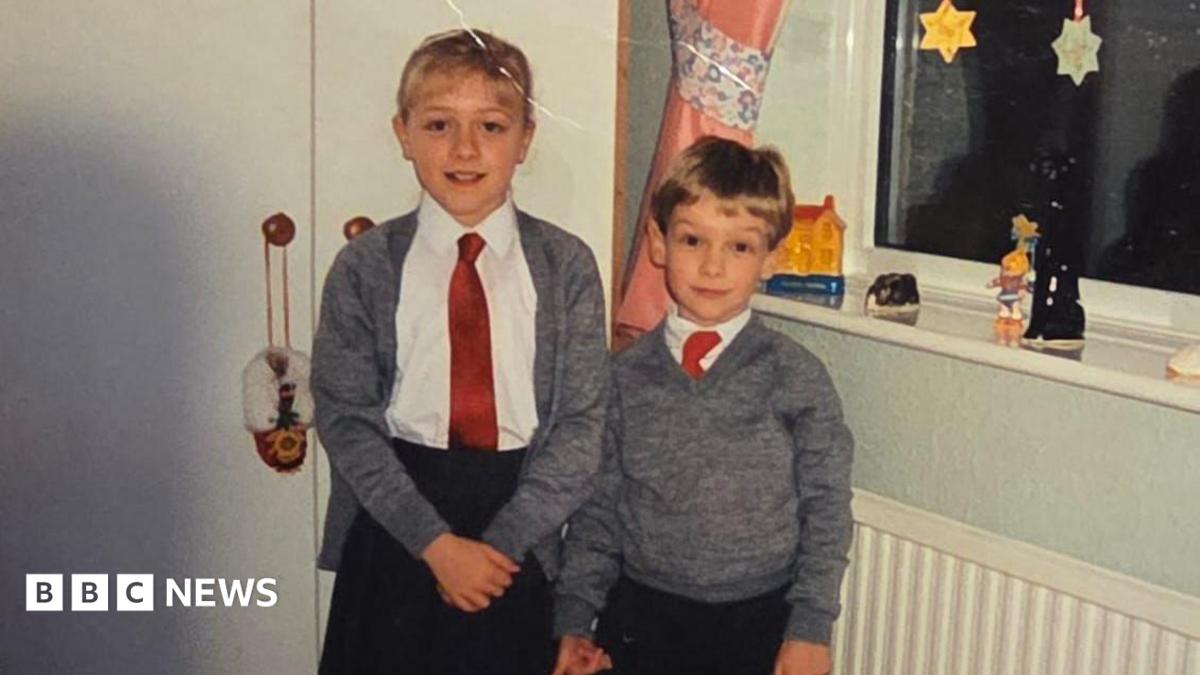 The childhood photo of Claire and Peter which was sent to Dalibor Sentic in the care package he recieved in 1992. Claire and Peter are photographed in school uniforms, stood in what appears to be a bedroom. Claire is taller than Peter and stood to the left of the image. She is wearing a black skirt, white blouse, red tie, and grey cardigan. She has blonde hair with a fringe. Her hair is tied up. Peter is shorter than Claire and is photographed to the right of her. He is wearing black trousers, a white shirt and red tie, with a grey jumper over the top.