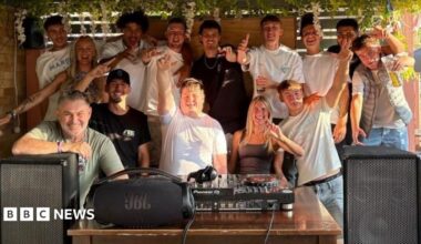Adam, centre, is surrounded by a group of people behind DJ decks and large speakers. Everyone is smiling with their hands in the air.