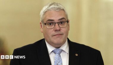 DUP leader Gavin Robinson, with short grey hair, wearing glasses, a white shirt, blue tie and black jacket.