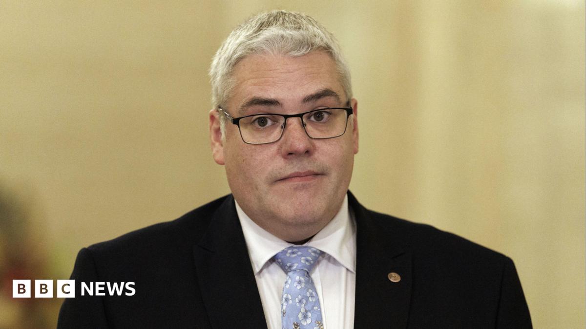 DUP leader Gavin Robinson, with short grey hair, wearing glasses, a white shirt, blue tie and black jacket.