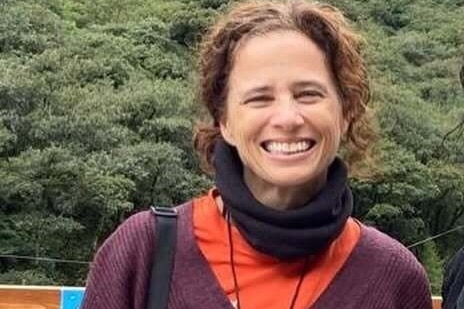A smiling, middle-aged woman with dark, curly hair standing in front of an expanse of bushland.