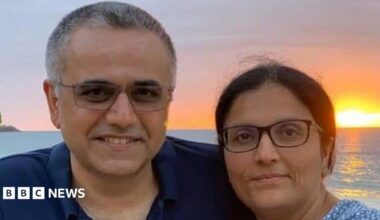 A middle aged man and woman, both wearing glasses, looking at the camera. They're smiling. There's a sunset over the sea in the background.