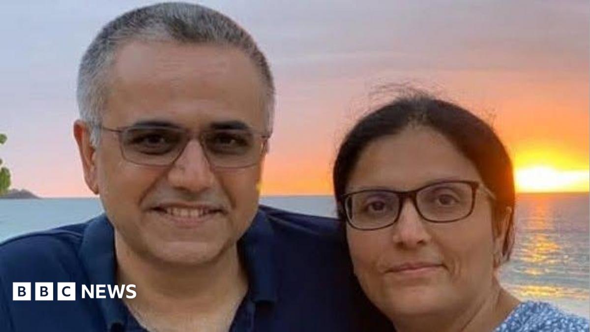 A middle aged man and woman, both wearing glasses, looking at the camera. They're smiling. There's a sunset over the sea in the background.