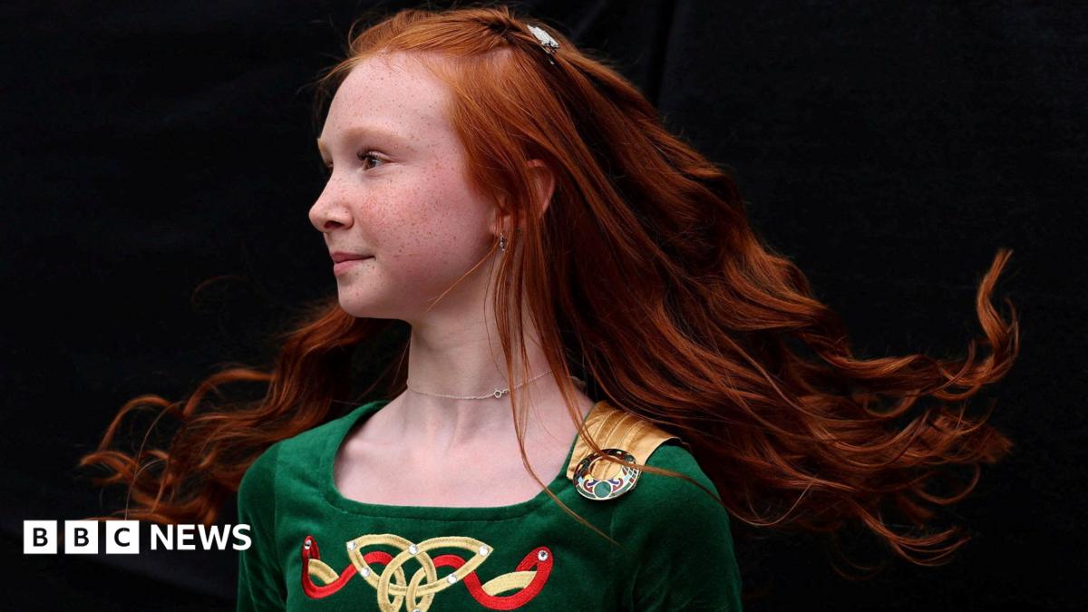 A group of young girls play instruments on a stage including a guitar, a fiddle, and tin whistles.  They are standing on a stage in front of an Irish language sign which says Fleadh Cheoil na hÉireann.