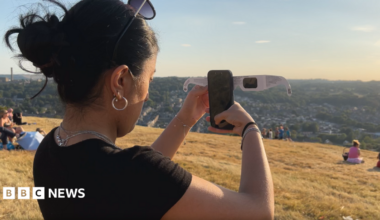 A woman holds her phone up to some solar glasses while standing on a hill overlooking Bradford.