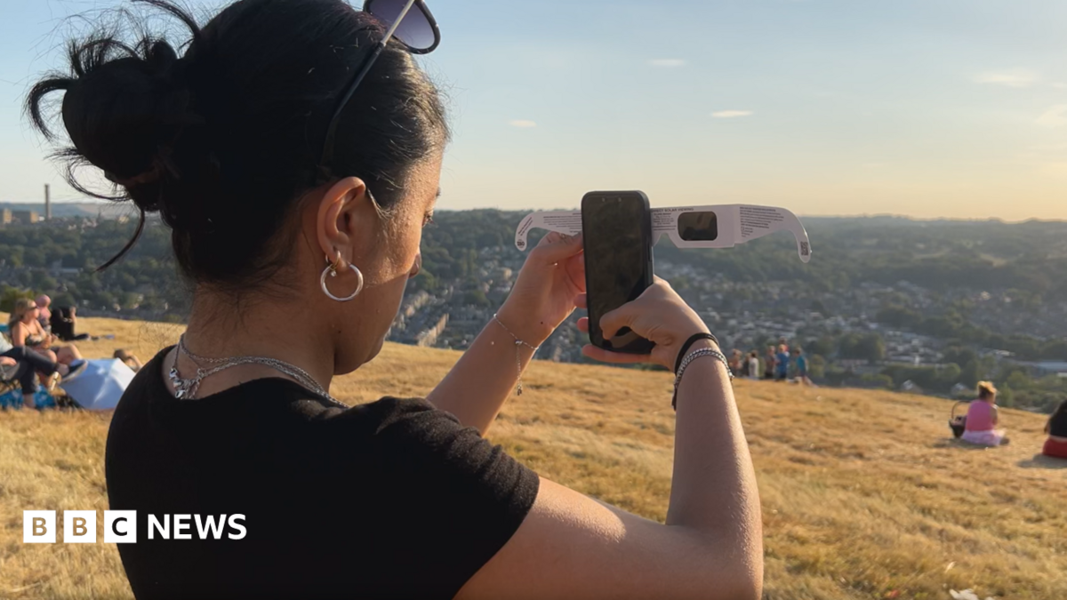 A woman holds her phone up to some solar glasses while standing on a hill overlooking Bradford.