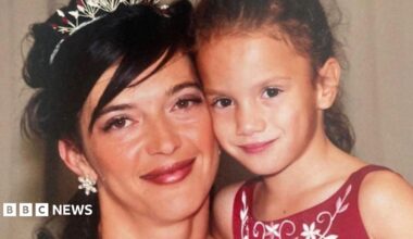 A woman with dark hair tied up with a silver tiara looks at the camera and cuddles a young girl with tied back dark hair and a red dress