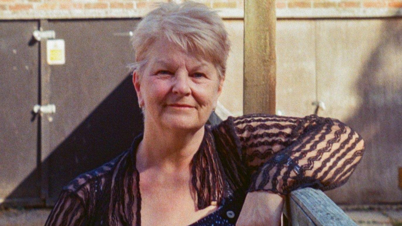 Shirley smiles as she leans on a wooden gate behind two garages. She has short, light blonde hair and wears a sheer shirts with a black wiggly pattern on it.