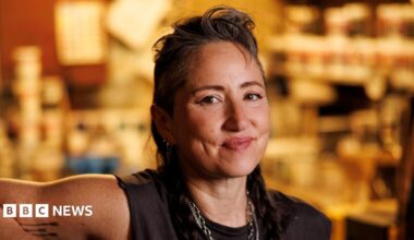 KT Tunstall in black T-shirt and necklaces, with her hair swept back off her face. She is smiling. The gold-coloured backdrop is out of focus.