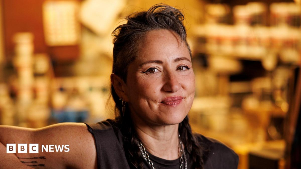 KT Tunstall in black T-shirt and necklaces, with her hair swept back off her face. She is smiling. The gold-coloured backdrop is out of focus.