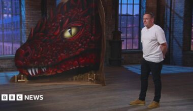 Nathan Murdoch is standing in a studio wearing a white T-shirt and dark trousers. There is a large painting of a scaly, red dragon with yellow eyes next to him.