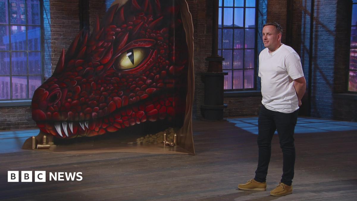 Nathan Murdoch is standing in a studio wearing a white T-shirt and dark trousers. There is a large painting of a scaly, red dragon with yellow eyes next to him.
