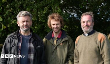 Three men sending side by side. First man has short grey hair and is wearing a blue t shirt, checked shirt and black jacket. The second man is wearing a khaki coloured jacket and the third man - a reverend - is wearing a khaki coloured jumper