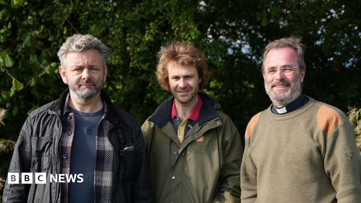 Three men sending side by side. First man has short grey hair and is wearing a blue t shirt, checked shirt and black jacket. The second man is wearing a khaki coloured jacket and the third man - a reverend - is wearing a khaki coloured jumper