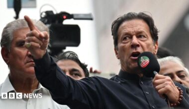 Imran Khan wearing a black button-up shirt and speaking into a microphone, points dramatically. Behind him are other men, and a television camera