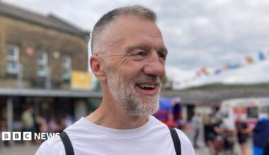 A man with greying short hair and a beard stands laughing in the street.