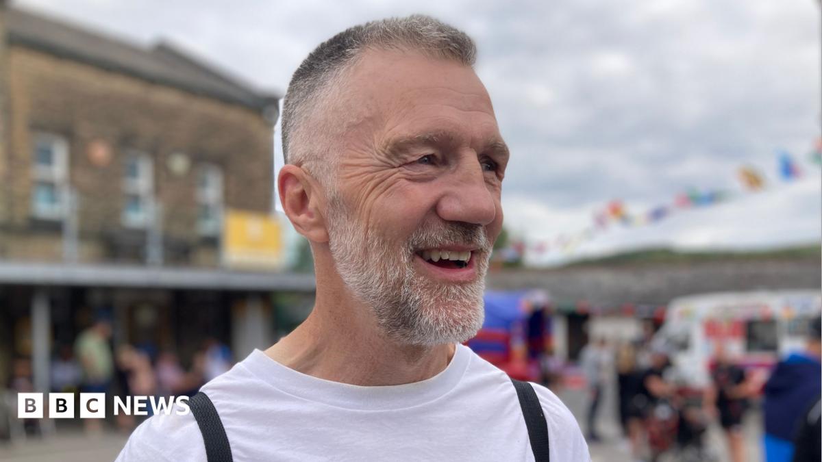 A man with greying short hair and a beard stands laughing in the street.