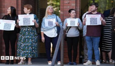 Four women and a man are standing in front of a brick building holding small white posters that say 'regulate our funeral industry'