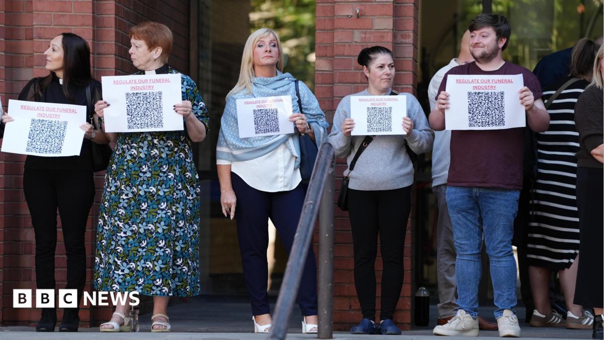 Four women and a man are standing in front of a brick building holding small white posters that say 'regulate our funeral industry'