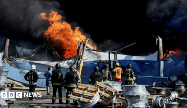 Wide shot showing Ukrainian firefighters in black and yellow uniforms observing flames rising from the debris of what appears to be a warehouse following a drone strike.