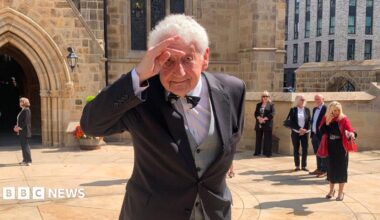 A man with grey hair makes a hand salute to his head outside a cathedral