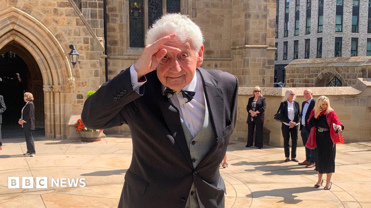 A man with grey hair makes a hand salute to his head outside a cathedral