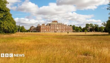 A large stately home is in the distance. A large field which has dry grass and hay leads to the home which is three storeys. Large green trees are surrounding the building.