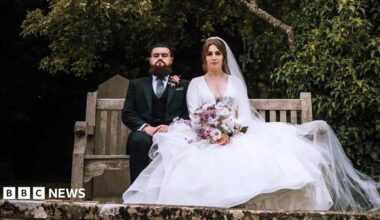 A bride and groom sit side-by-side on a wooden bench looking at the camera, with expressionless faces. The groom is wearing a dark suit with a pink flower attached to his jacket. The bride is wearing a long white dress draped over the side of the bench, and is holding a bouquet of flowers.