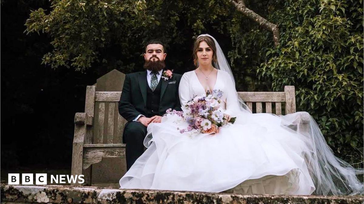 A bride and groom sit side-by-side on a wooden bench looking at the camera, with expressionless faces. The groom is wearing a dark suit with a pink flower attached to his jacket. The bride is wearing a long white dress draped over the side of the bench, and is holding a bouquet of flowers.
