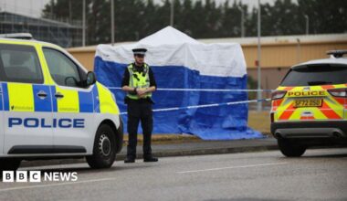 A police officer standing in front of a police cordon and blue forensic tent and two police vehicles in Arbroath.