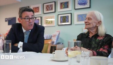 Andy Burnham sits at a table in a care home, talking face-to-face with a resident. The prime minister is in a dark suit and glasses and leans forward while speaking, and the other, with long white hair, sits opposite. Several glasses of water and cups are on the table, with framed artwork displayed on the wall behind them.
