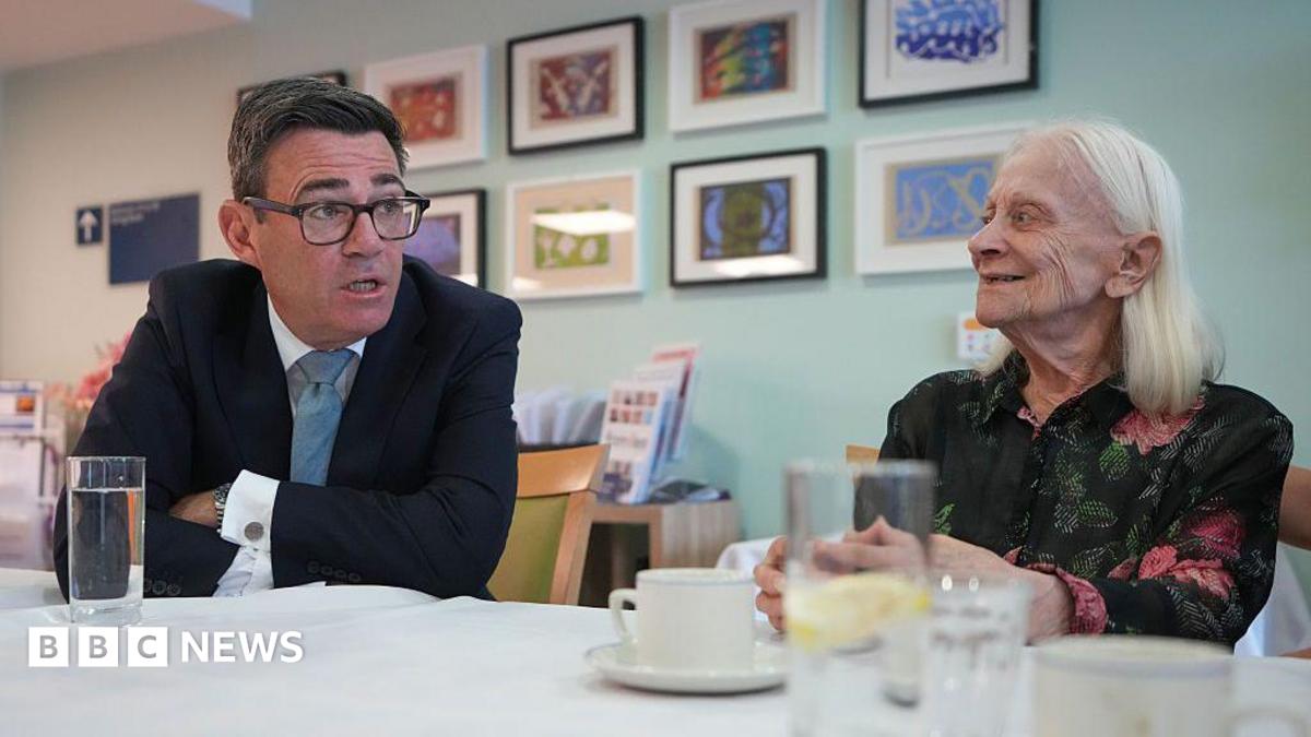 Andy Burnham sits at a table in a care home, talking face-to-face with a resident. The prime minister is in a dark suit and glasses and leans forward while speaking, and the other, with long white hair, sits opposite. Several glasses of water and cups are on the table, with framed artwork displayed on the wall behind them.