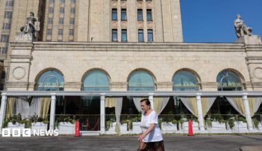 A woman wearing a red jumper and sunglasses, as well as a little bag on her right shoulder, talks on the phone in front of an exterior view of the Central Bank of the Russian Federation building on top of which the Russian flag can be seen