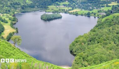 Grasmere, which is a lake with a small island towards the back left. The island is covered in trees and the water on the lake is calm. Bright green fields and trees surround the perimeter of the lake. Houses and buildings are scattered across the landscape.