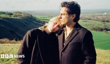 An image of a blonde woman who is wearing all black leaning on a man with brown hair who is also wearing all black in the middle of a field.