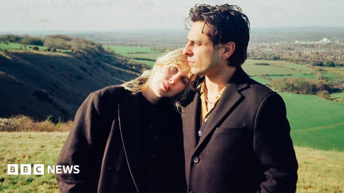 An image of a blonde woman who is wearing all black leaning on a man with brown hair who is also wearing all black in the middle of a field.